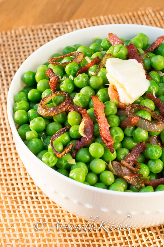 White bowl filled with green peas mixed with bacon and shallots. Topped with a pat of butter.