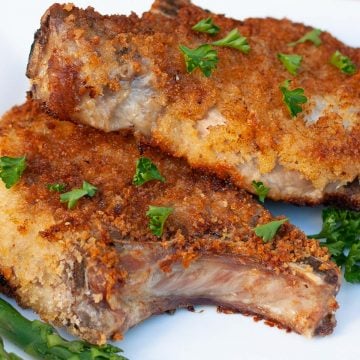 Pan fried breaded pork chops on white plate garnished with parsley flakes.
