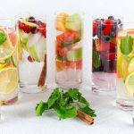 Colorful infused water glasses lined up, each filled with assorted fruits like kiwi, berries, citrus, and herbs like mint.