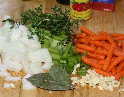 Chopped onions, celery, carrots, and garlic with bay leaves and thyme on wood cutting board.