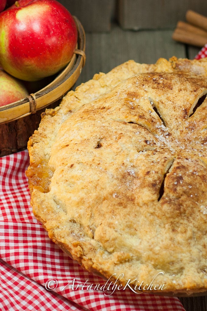 Apple pie with golden flaky crust on checkered cloth with basket of apples in background.