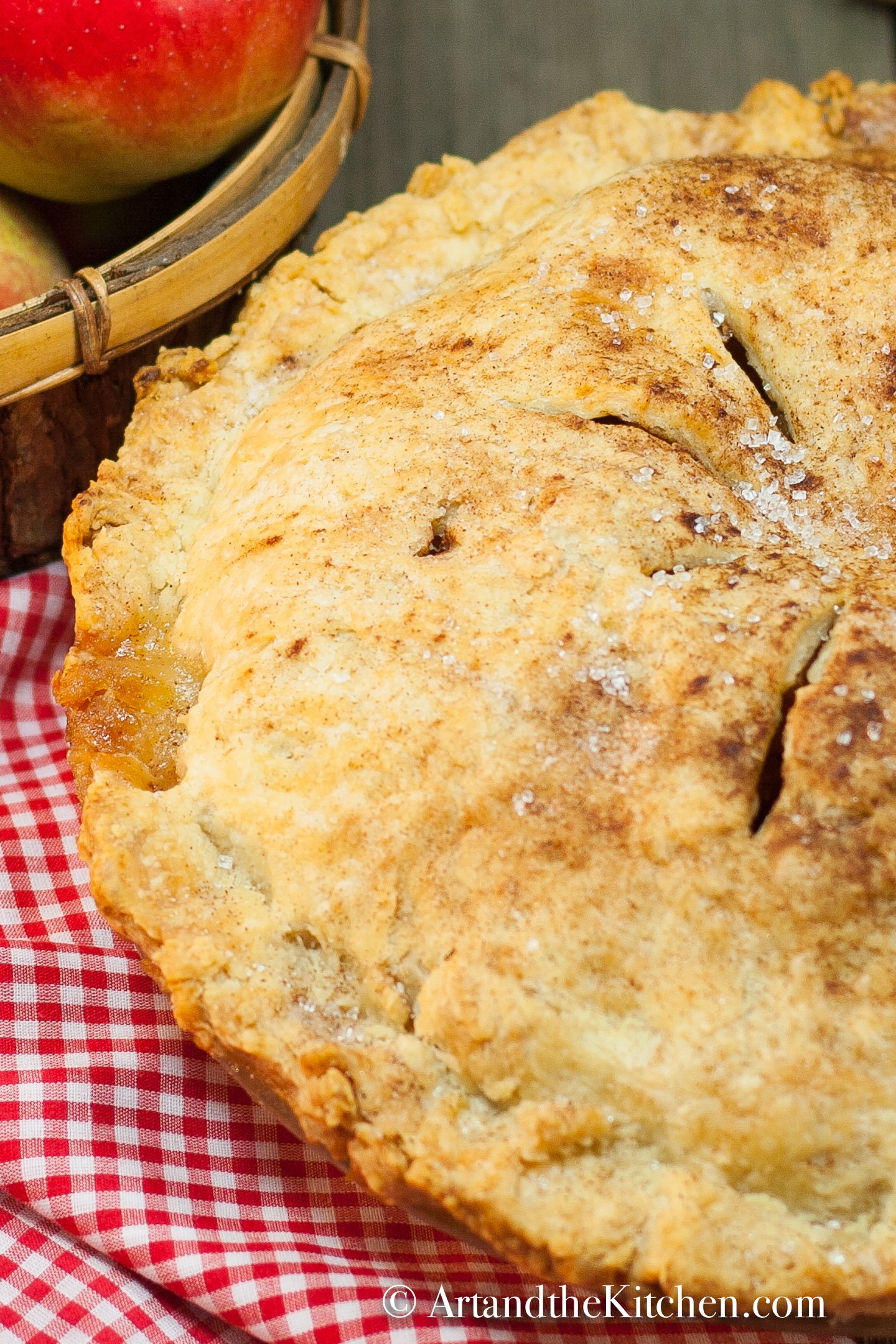 Apple pie with golden flaky crust on checkered cloth with cinnamon sticks and basket of apples in background.