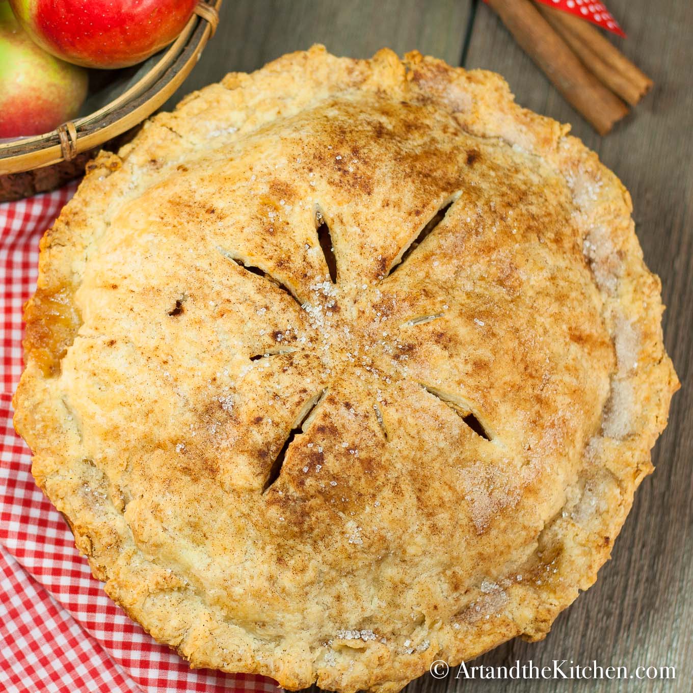 Apple pie with golden flaky crust on checkered cloth with cinnamon sticks and basket of apples in background.