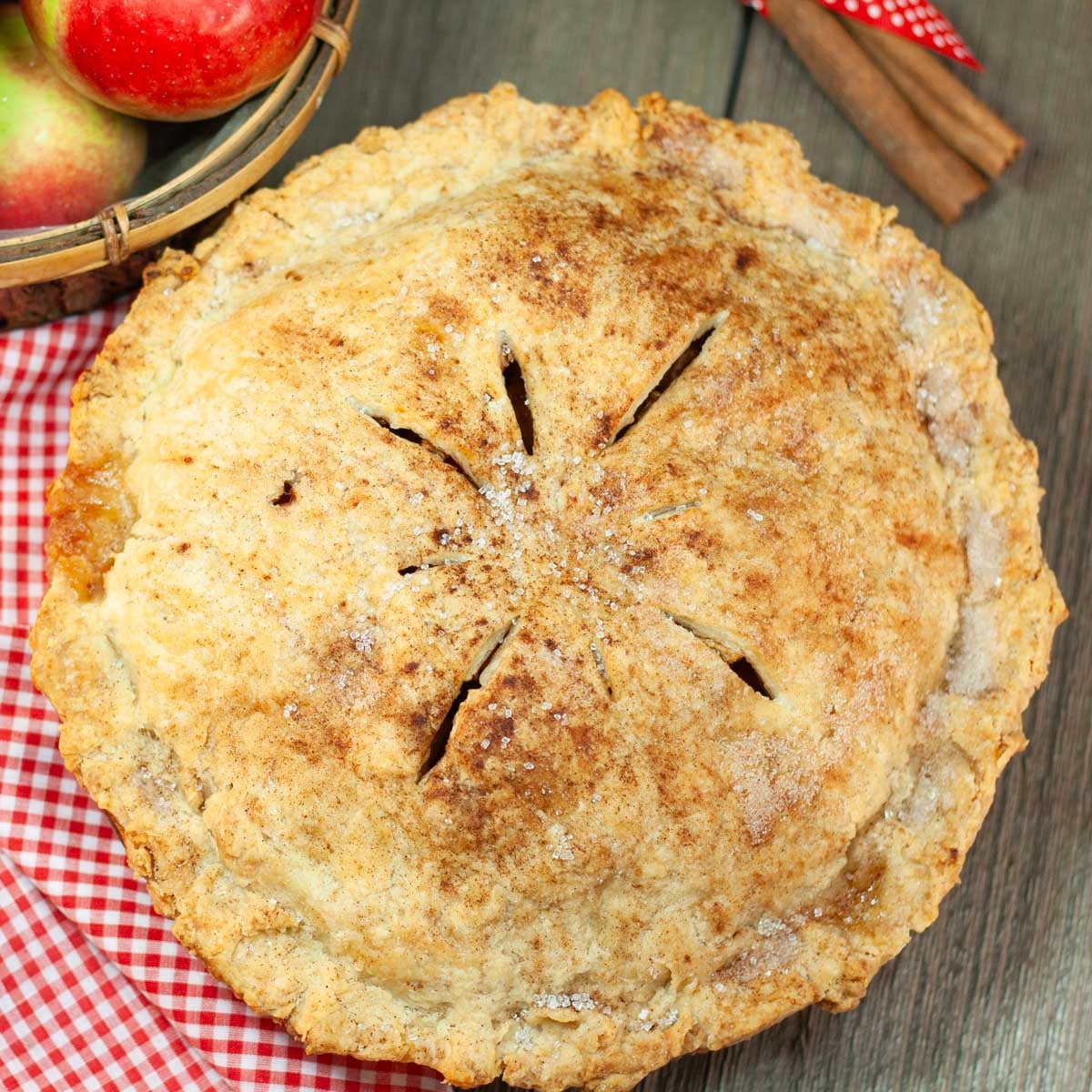 Apple pie with golden flaky crust on checkered cloth with cinnamon sticks and basket of apples in background.