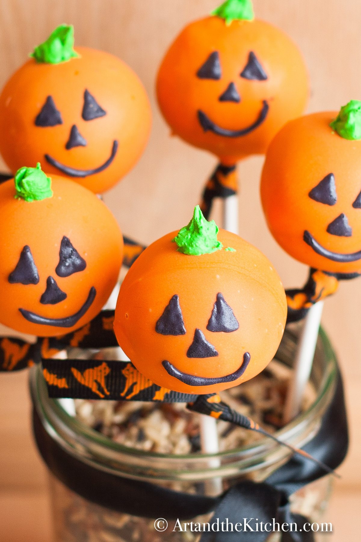 Five orange colored cake pops decorated like jack o lanterns displayed in a decorative jar.