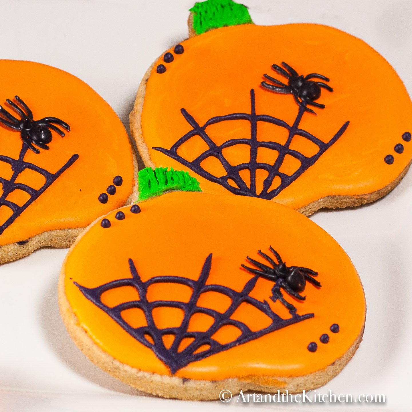 Three pumpkin shaped, orange frosted sugar cookies on a white plate. Decorated with black spider and web icing.