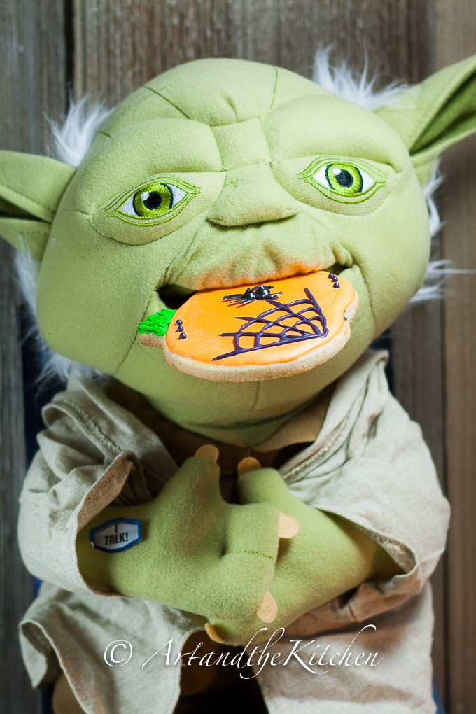 Plush Yoda doll eating an orange sugar cookie that is decorated with a spider web.