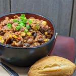 A black bowl filled with chili, corn, and beans, garnished with chopped green onions, served with a bread roll.