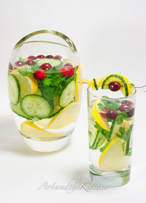 Pitcher and tall glass of water and cucumber, lemon slices, parsley and cilantro.