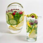 Pitcher and tall glass of water and cucumber, lemon slices, parsley and cilantro and cranberries.