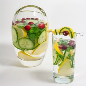 Pitcher and tall glass of water and cucumber, lemon slices, parsley and cilantro and cranberries.