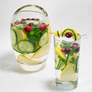 Pitcher and tall glass of water and cucumber, lemon slices, parsley and cilantro and cranberries.