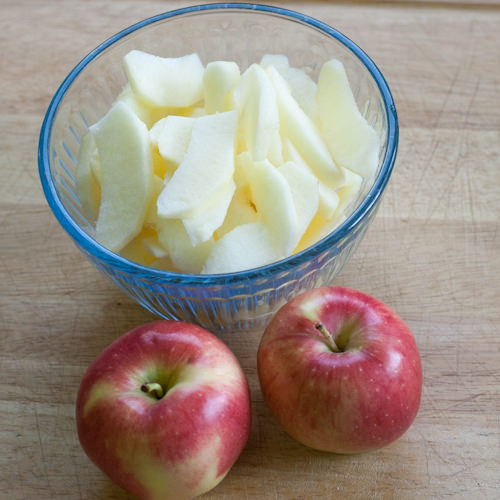 Glass bowl filled with apples slice beside two whole apples.