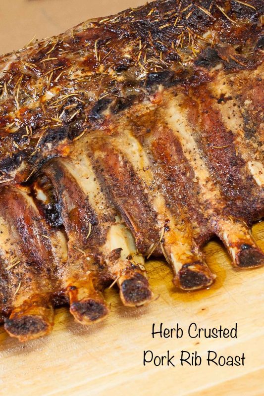 Cooked whole pork rib roast topped with herbs on wood cutting board.