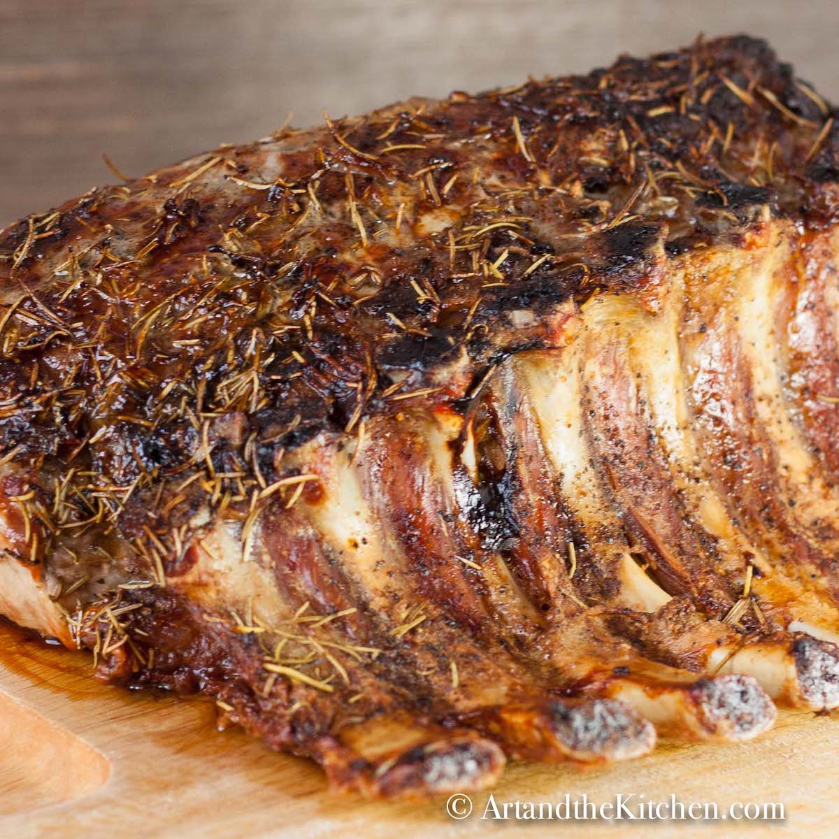 Cooked whole pork rib roast topped with herbs on wood cutting board.