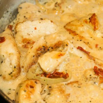 Chunks of chicken with shallots and sun-dried tomatoes in a creamy sauce simmering in a stainless steel skillet.