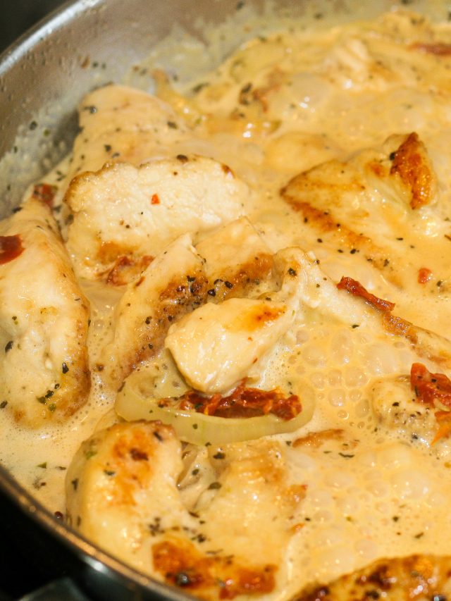 Chunks of chicken with shallots and sun-dried tomatoes in a creamy sauce simmering in a stainless steel skillet.