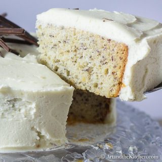 Slice of banana cake with layer of cream cheese frosting lifted from whole cake with cake server on a decorative glass plate.