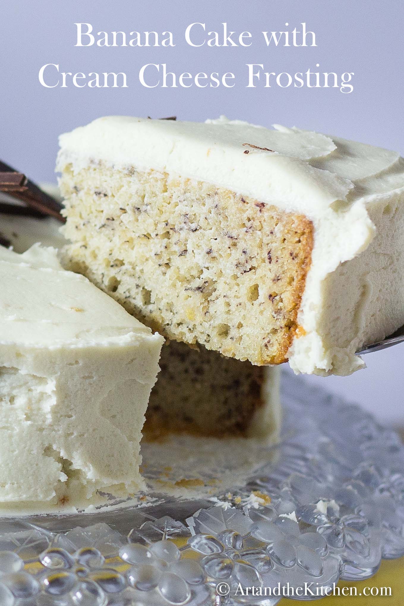 Slice of banana cake with layer of cream cheese frosting lifted from whole cake with cake server on a decorative glass plate.