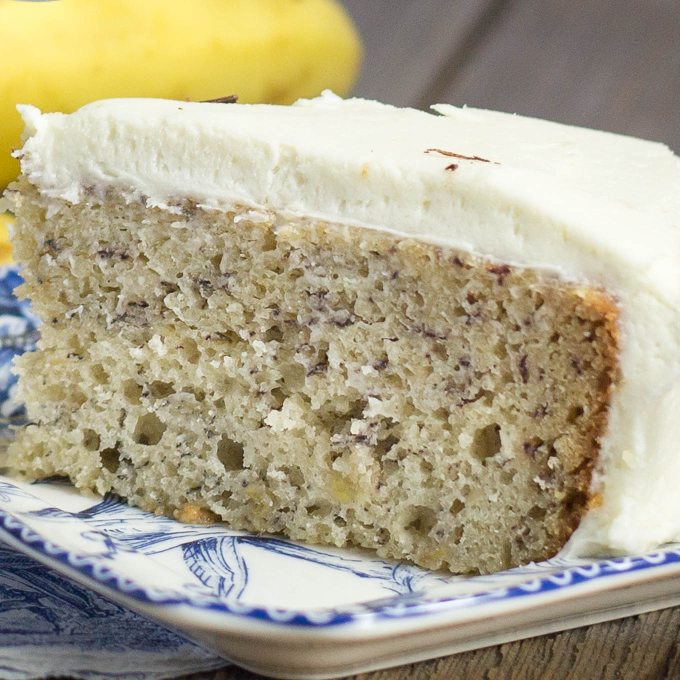 Slice of banana cake with layer of cream cheese frosting on a decorative blue plate.