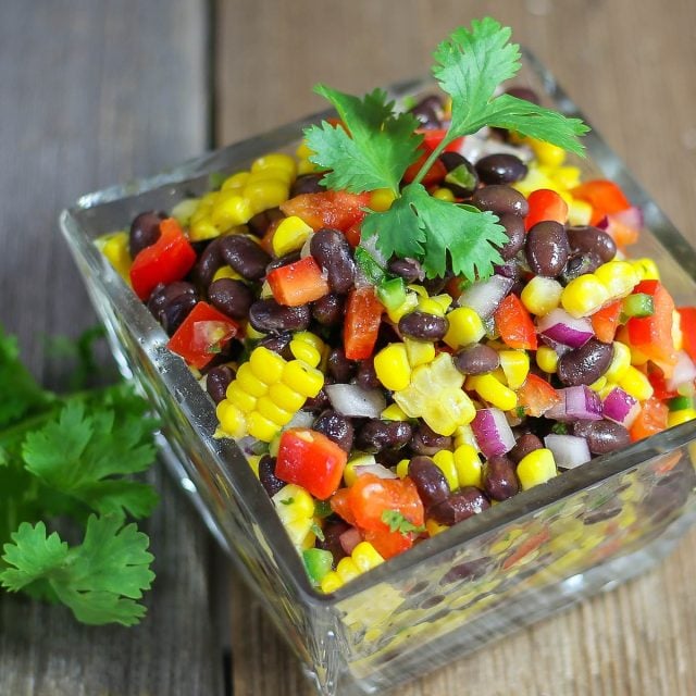 A vibrant salad in a glass dish features black beans, corn, red bell peppers, and chopped onions, garnished with fresh cilantro.