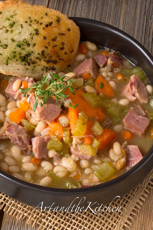 Ham and bean soup with chunks of ham, carrots and celery served in a black bowl and garnished with garlic toast on side of bowl