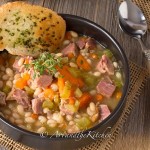 bowl of ham and bean soup with chunks of ham, navy beans, carrots and celery. Garlic bread on side of bowl, garnishes with thyme sprig.
