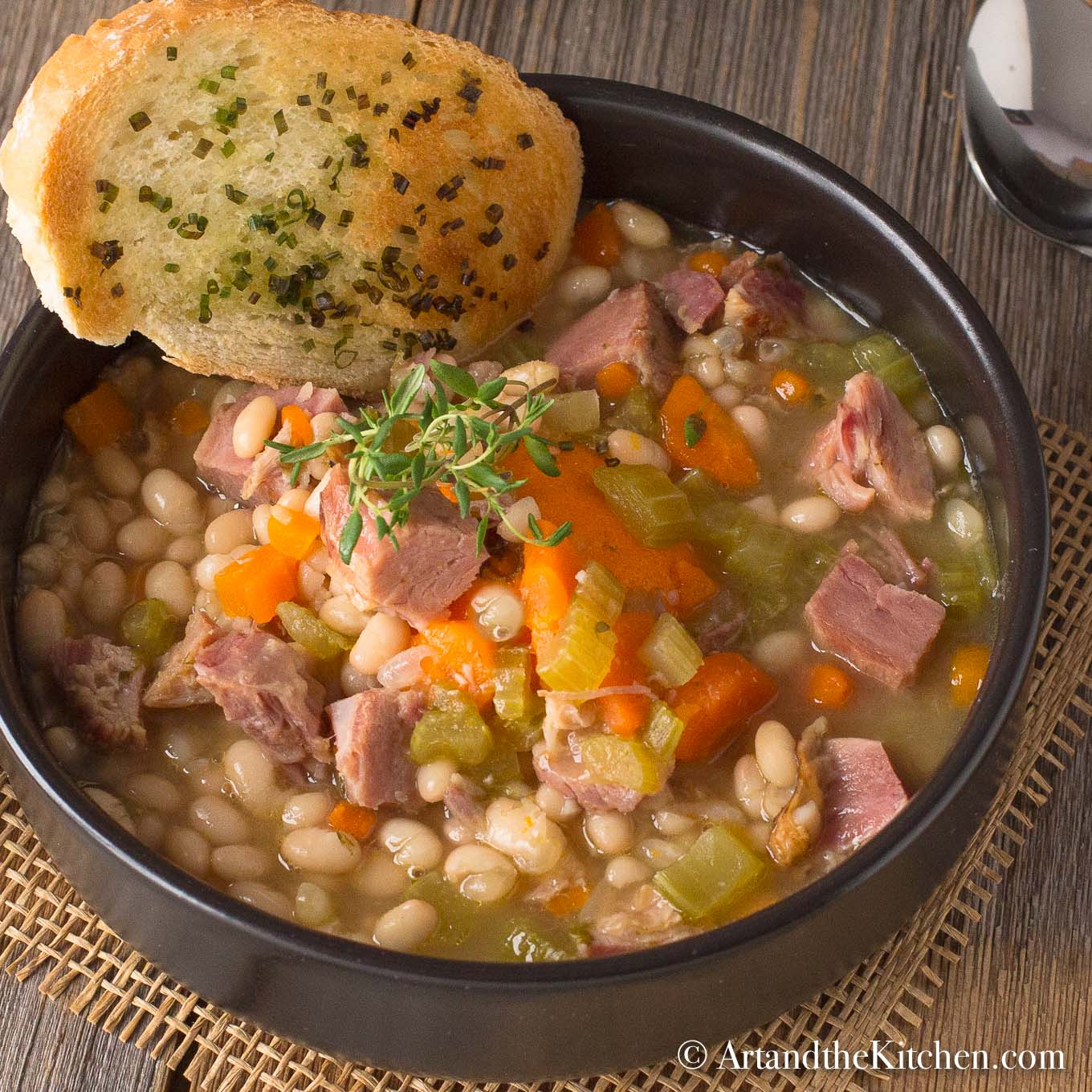 Black bowl filled with chunky soup of ham and beans with piece of garlic toast.