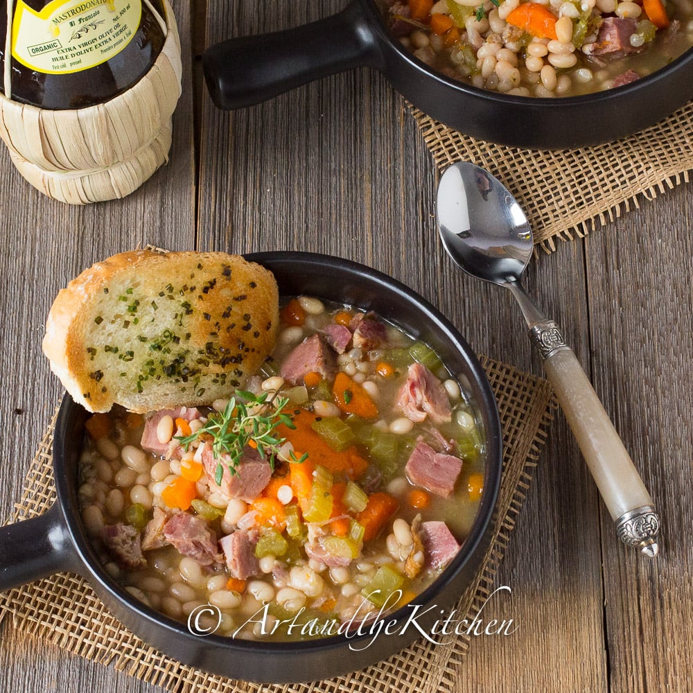 Black bowl filled with ham and bean soup with garlic toast on side. Decorative spoon and wine bottle in background.