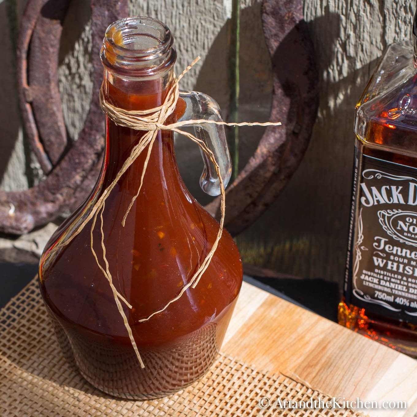 Decorative glass jar filled with homemade BBQ sauce on wood board with bottle of Jack Daniel's whiskey in background.