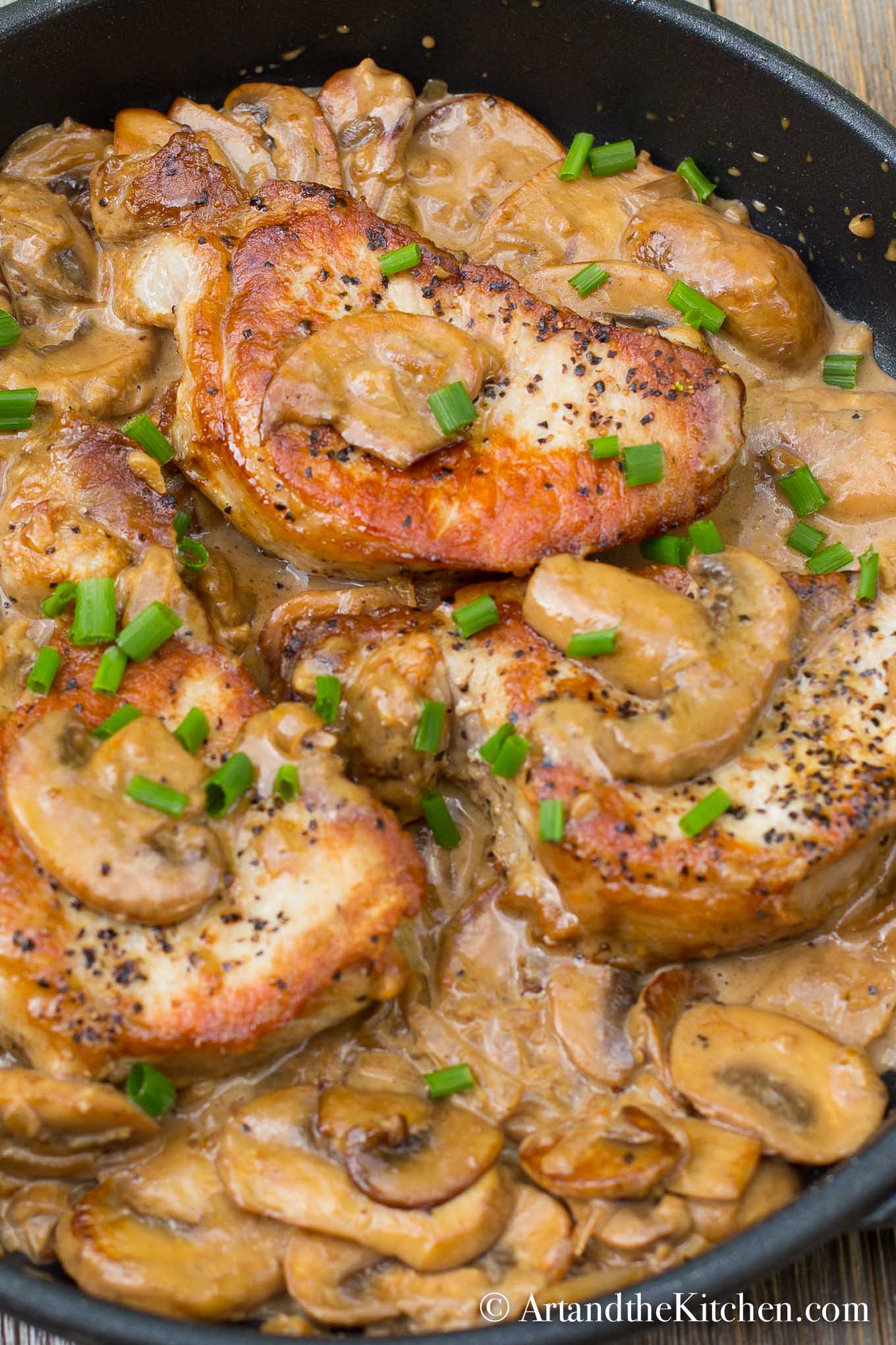 Cast iron frying pan with seared pork chops in a creamy mushroom sauce, garnished with green onion slices. 