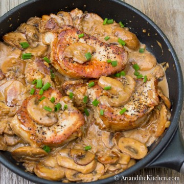 Cast iron frying pan with seared pork chops in a creamy mushroom sauce, garnished with green onion slices.