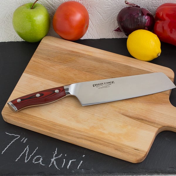 Nakiri knife on wood cutting board with fruit and vegetables in background.