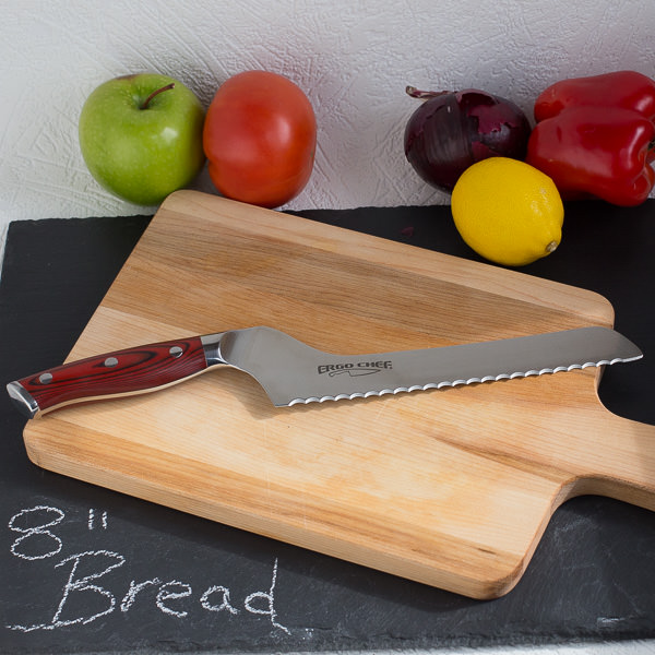 Bread knife on wood cutting board with fruit and vegetables in background.