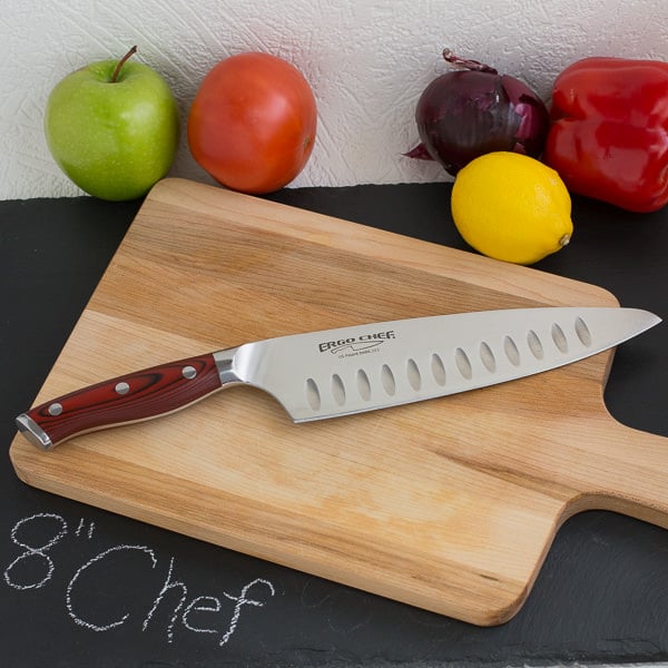 Chef knife on wood cutting board with fruit and vegetables in background.