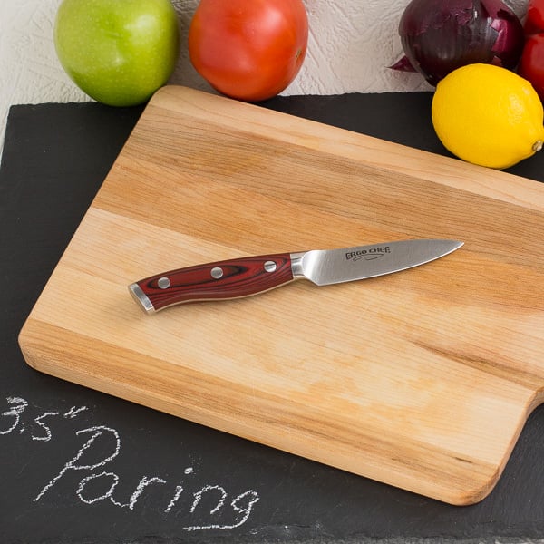 Paring knife on wood cutting board with fruit and vegetables in background.