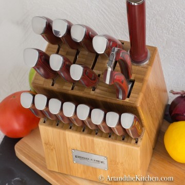 Wood knife block filled with set of knives on wood cutting board with fruit and vegetables in background.
