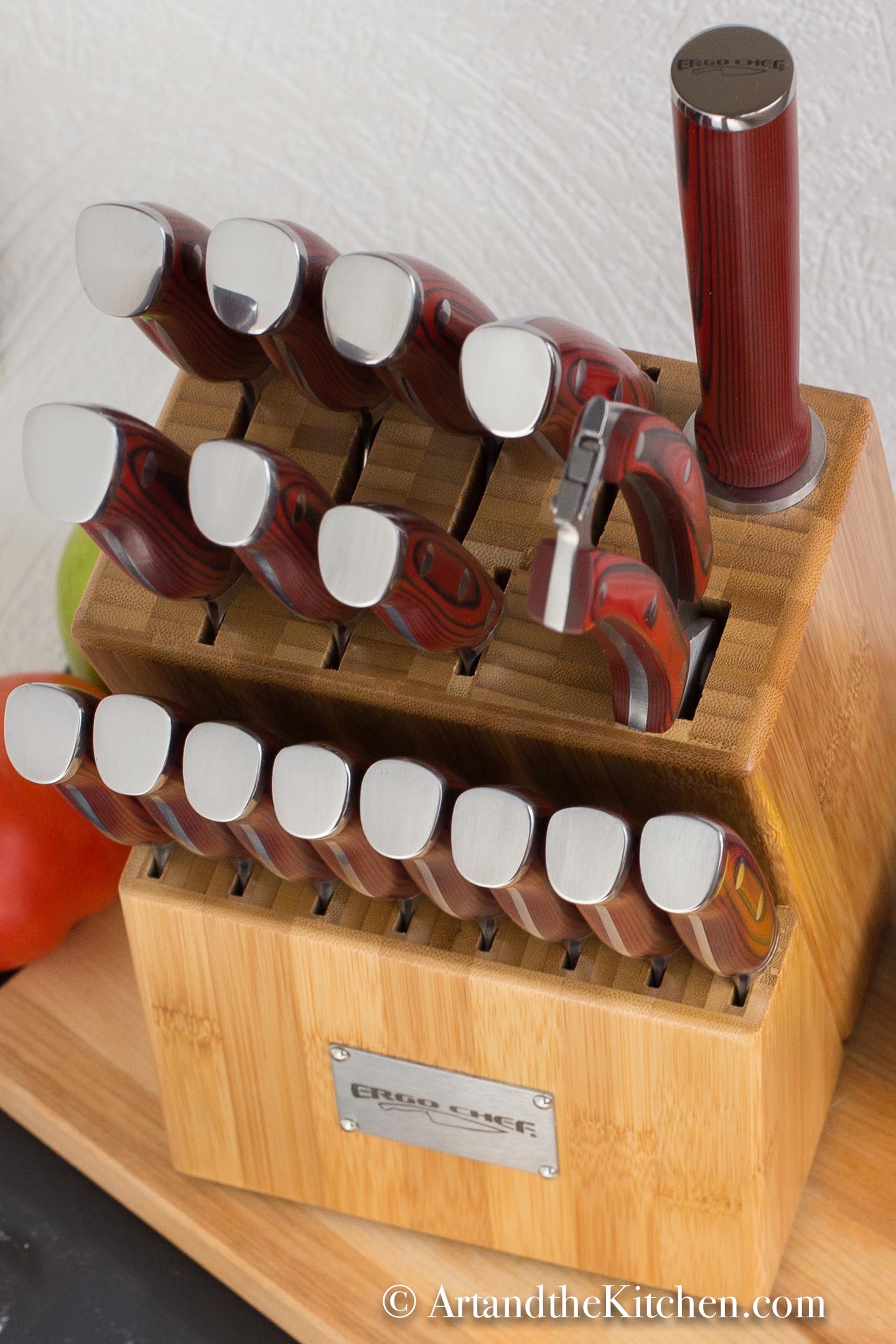 Wood knife block filled with set of knives on wood cutting board with fruit and vegetables in background.