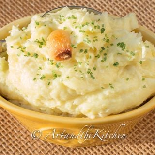 Yellow bowl filled with mashed potatoes, topped with clove of roasted garlic.