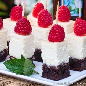 Bite size pieces of cheesecake with brownie crust topped with a raspberry held with wood toothpick