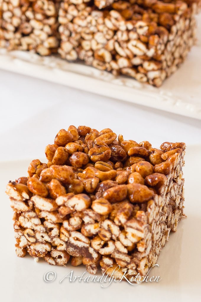 Single piece of square make with puffed wheat and gooey syrup mixture with plate of squares in background on white plate.