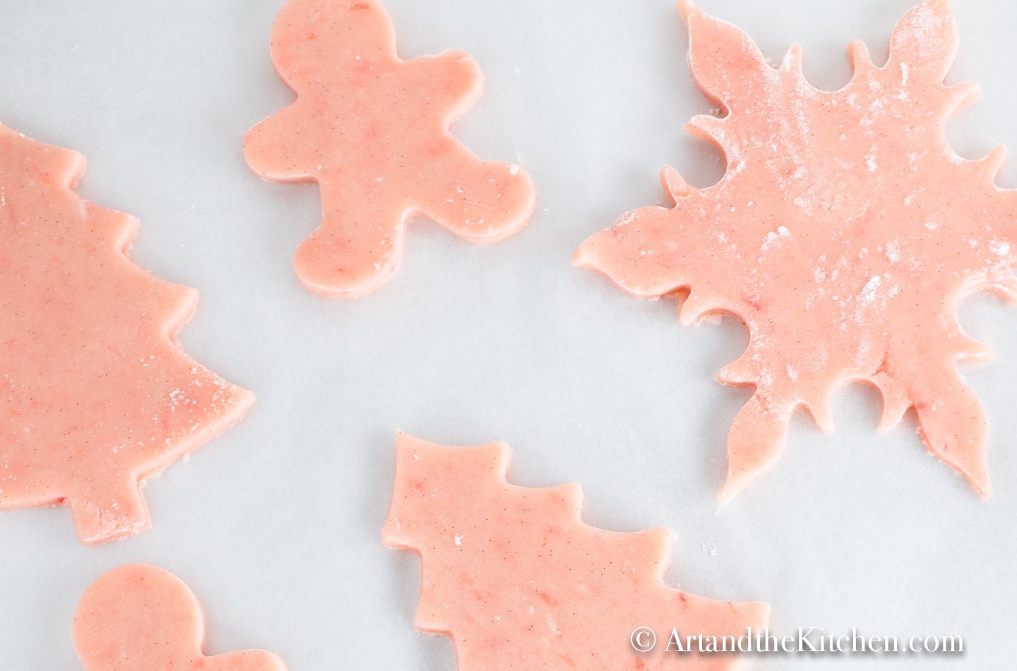 Pink sugar cookie dough cut into Christmas cookie shapes on baking sheet lined with white parchment paper.