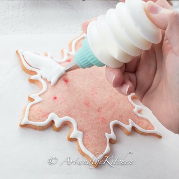 Pink snowflake shaped cookie being decorated with royal icing from squeeze bottle.