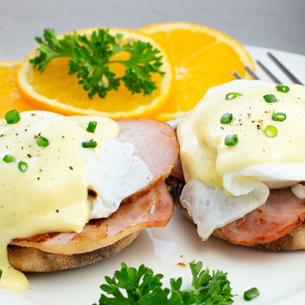 Close-up of Eggs Benedict with Canadian bacon on toasted muffins, topped with Hollandaise sauce, garnished with chopped chives. Orange slices and parsley on the side.