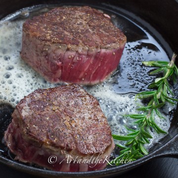 Two tenderloin steaks frying in cast iron skillet with rosemary sprig.