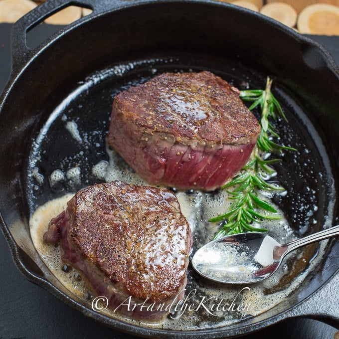 Two tenderloin steaks frying in cast iron skillet with rosemary sprig.