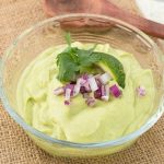 Avocado dip garnished with red onions, lime slice and cilantro in small glass bowl.