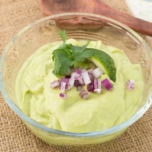 Avocado dip garnished with red onions, lime slice and cilantro in small glass bowl.