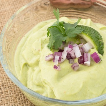 Avocado dip garnished with red onions, lime slice and cilantro in small glass bowl.