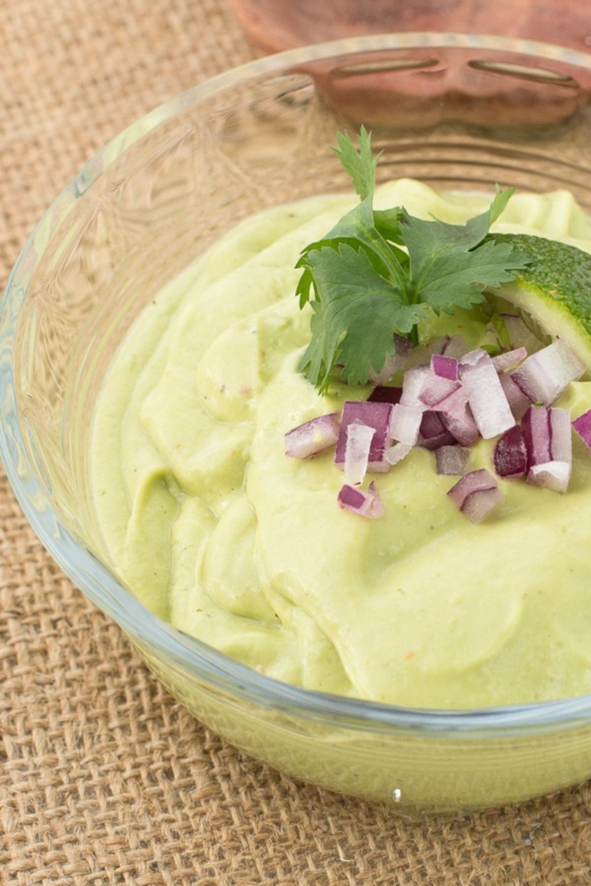 Avocado dip garnished with red onions, lime slice and cilantro in small glass bowl.